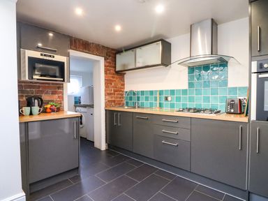 A kitchen with gray cabinets a microwave a kettle fruit on a counter turquoise tiles and a washing machine visible through a doorway at South Harbour Cottage in Weymouth