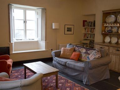 A living room with a wooden coffee table a sofa with cushions an armchair a window a floor lamp a bookcase and a wooden cabinet with plates at Thomas Cottage in Hartsop