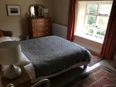 A bedroom with a bed covered with a blanket a wooden chest of drawers and a window with red curtains at Thomas Cottage in Hartsop