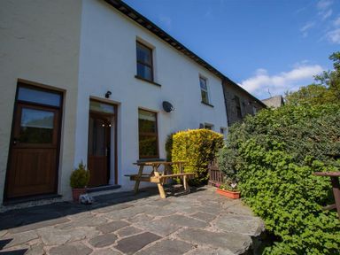 A paved patio with a wooden picnic table outside a white two-story house with wooden doors and windows and surrounding bushes at Robins Nest in Coniston