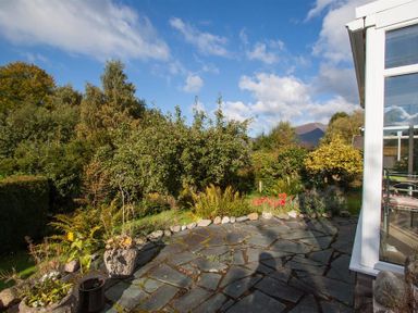 A stone patio with plants in pots and bushes surrounding it by a glass conservatory at Brimbles in Keswick