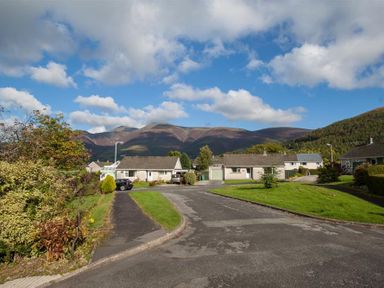 A residential street with bungalows and parked cars with mountains in the background at Brimbles in Keswick