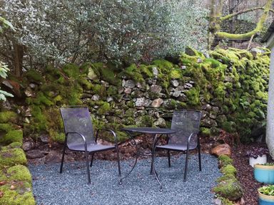 A small outdoor seating area with two metal chairs and a round table on gravel next to a mossy stone wall