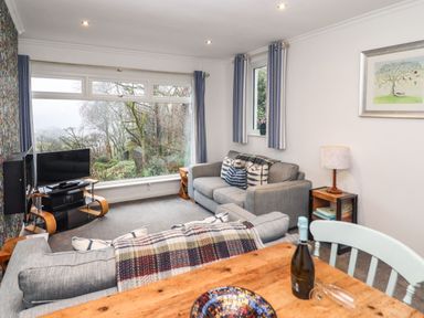 A living room with gray sofas a wooden table a television and large windows showing trees at Helm Crag in Grasmere