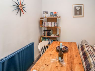 A dining area with a wooden table set with a bottle and glasses near a blue radiator and a wooden shelving unit with books and DVDs at Helm Crag in Grasmere