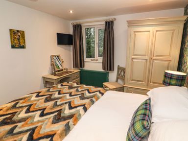 A bedroom with a bed covered in a chevron patterned blanket a wooden wardrobe a desk with a mirror and chair a window with curtains and a wall-mounted TV at Helm Crag in Grasmere