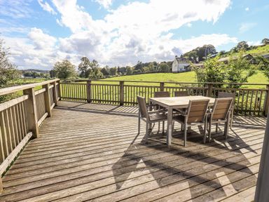 A wooden deck with a table and six chairs overlooking a green field with a house in the distance at How Beck in Hawkshead