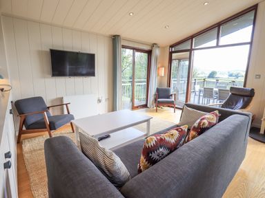 A living room with a gray sofa a white coffee table two armchairs a wall-mounted TV and large windows at How Beck in Hawkshead