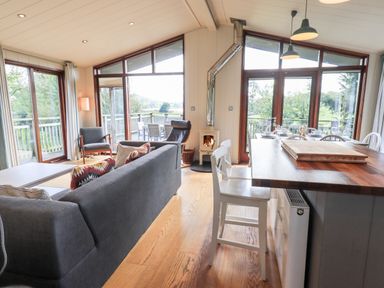 A living area with a gray sofa, chairs, dining table with chairs, wood burning stove, and large windows overlooking a deck at How Beck in Hawkshead