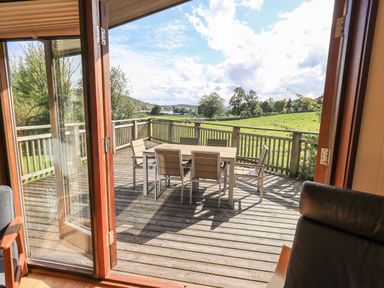 An outdoor wooden deck with a dining table and six chairs overlooking a green field at How Beck in Hawkshead