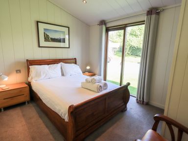 A bedroom with a wooden bed, bedside tables, lamps, rolled towels on the bed, a framed picture on the wall, and a sliding glass door at How Beck in Hawkshead