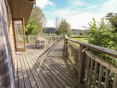 An outdoor wooden deck with a table and chairs overlooking trees and hills at How Beck in Hawkshead