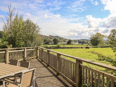A wooden deck with a table and chairs overlooking a grassy field with cows and trees at How Beck in Hawkshead