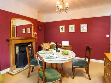 A dining room with a round table set with plates and wine glasses and four wooden chairs and a fireplace with a mirror above it at Greystones in Bowness