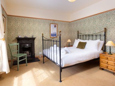 A bedroom with a metal bed frame white bedding a green chair a small black fireplace and wooden bedside tables with lamps at Greystones in Bowness