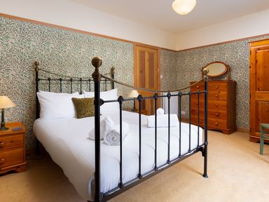 A bedroom with a black metal bed, wooden furniture, patterned wallpaper, and rolled white towels on the bed at Greystones in Bowness