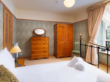A bedroom with a bed, wooden dresser with mirror, wardrobe, nightstand with lamp, chair, and window with curtains at Greystones in Bowness