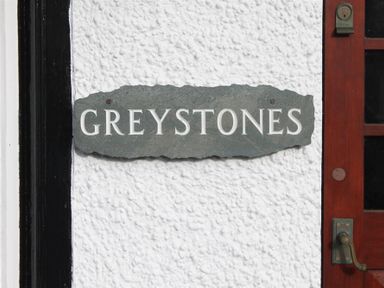 A slate house name sign reading GREYSTONES on a white textured wall next to a wooden door at Greystones Bowness