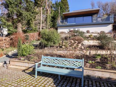 A blue bench on a paved area in front of terraced garden beds with bushes and a modern house in the background at Gillside in Millbeck