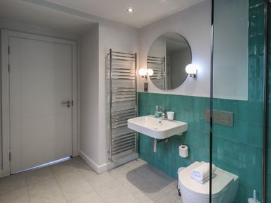 A bathroom with a white door tiled walls a wall-mounted sink a round mirror two wall lamps a heated towel rack and a toilet with folded towels at Gillside in Millbeck