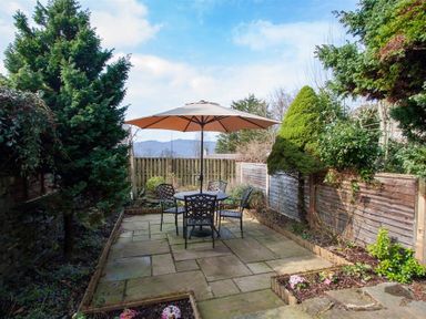 An outdoor patio with a table and four chairs under a large umbrella surrounded by trees and garden beds at Swallow's Nest in Windermere