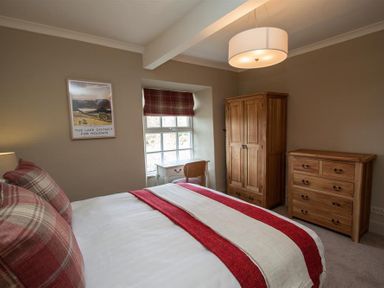 A bedroom with a bed a wooden wardrobe a wooden chest of drawers a small desk and chair and a window with plaid blinds at Swallow's Nest in Windermere