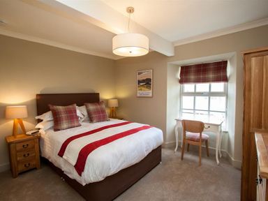 A bedroom with a double bed with pillows and a red striped bed runner with bedside tables and lamps and a window with red plaid blind and a desk and chair at Swallow's Nest in Windermere