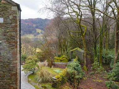 A stone pathway with bushes and mossy trees next to a stone building and a small stone structure in a wooded area at Silver Howe in Grasmere