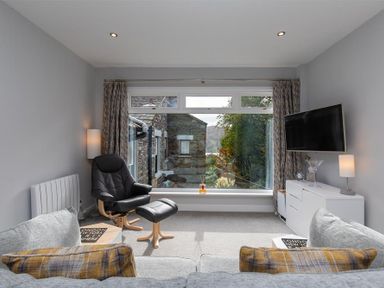 A living room with a large window showing an outside view a black armchair and footstool a gray sofa with cushions a white sideboard a wall-mounted television and table lamps at Silver Howe in Grasmere