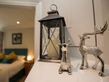 A candle lantern and two silver deer figurines on a white surface with a bed and nightstand in the background at Silver Howe in Grasmere