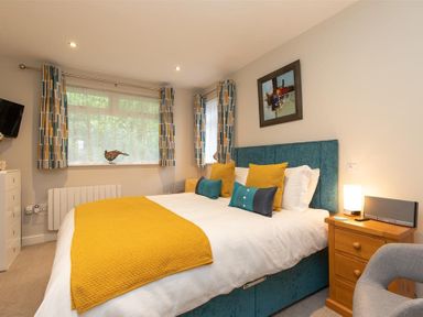 A bedroom with a bed and yellow and teal pillows a wooden bedside table a gray chair and windows with patterned curtains at Silver Howe in Grasmere
