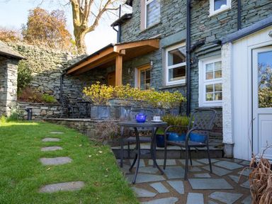 An outdoor patio with a small round table and two metal chairs on a stone floor near a stone house with potted plants along a raised area at Briardale Cottage in Ambleside