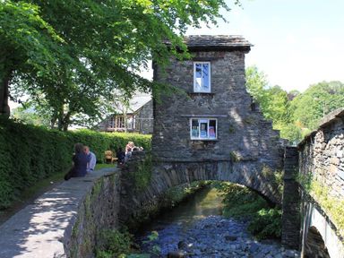A stone bridge over a small stream with a stone building on top and people sitting on a stone wall beside it at Briardale Cottage in Ambleside