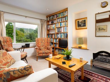 A living room with patterned armchairs a wooden coffee table a bookshelf and a window showing trees and hills at The Hayloft in Ambleside