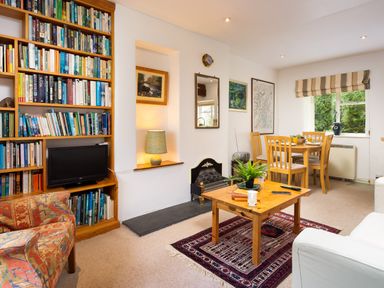 A living room with a wooden bookshelf filled with books a small TV a wooden coffee table with plants and books a dining table with chairs and windows at The Hayloft in Ambleside