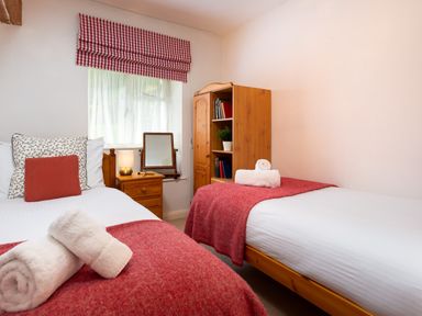 A bedroom with two single beds with red blankets and white towels a wooden nightstand with lamp and mirror a wooden bookshelf and a window with checked curtain at The Hayloft in Ambleside