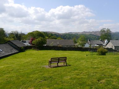A wooden bench on a grass field with stone walls and houses with tiled roofs in the background at Falls View Cottage in Ambleside