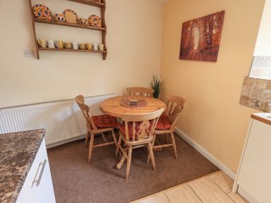 A kitchen dining area with a round wooden table and four chairs with cushions a wall shelf with plates and mugs and a framed picture on the wall at Hideaway Cottage in Ambleside