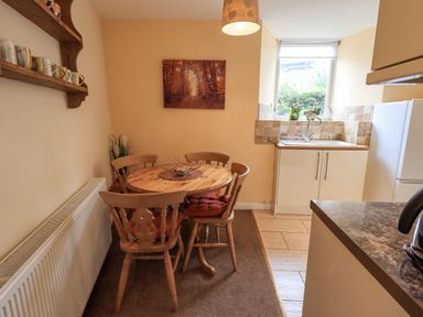 A kitchen area with a round wooden dining table and chairs a sink under a window and a shelf with mugs at Hideaway Cottage in Ambleside