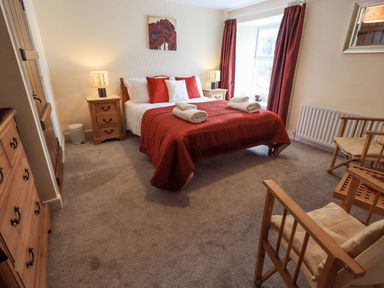 A bedroom with a double bed red bedding two bedside tables with lamps a wooden dresser two wooden chairs and a window with red curtains at Hideaway Cottage Ambleside