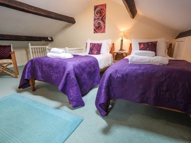 A bedroom with two single beds covered with purple bedspreads and white towels placed on them a wooden nightstand with a lamp between the beds and a chair in the corner at Hideaway Cottage Ambleside