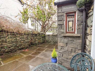A small stone patio with a metal table and chair a potted plant and a stone wall with a tree behind at Hideaway Cottage in Ambleside