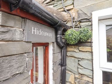 Stone wall with a black sign reading Hideaway black drainpipe and two hanging green plants at Hideaway Cottage in Ambleside
