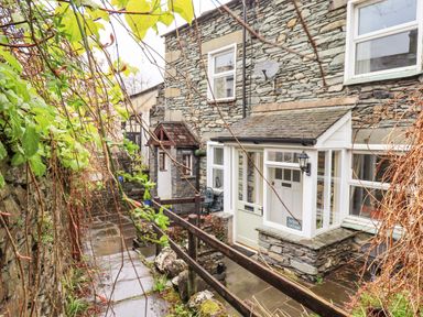 A stone exterior path leading to the entrance of a cottage with plants and a wooden fence at Hideaway Cottage in Ambleside