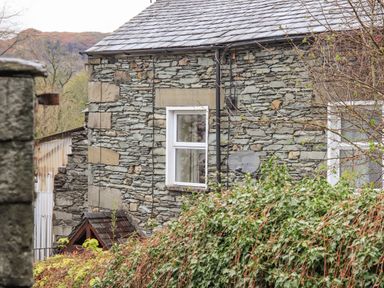 A stone house with two white framed windows and a satellite dish with greenery in front at Hideaway Cottage Ambleside