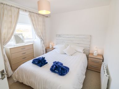 A bedroom with a double bed with white bedding and blue robes on it two wooden bedside tables a wooden chest of drawers and a window with curtains at Bryn Mor Church Bay