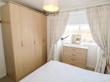 A bedroom with a wooden wardrobe, chest of drawers, window with curtains, and a bed with white bedding at Bryn Mor in Church Bay