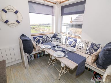 A dining nook with wooden bench seating, cushions with marine designs, a wooden table set with plates and glasses, two windows with blinds and a life ring on the wall at Bryn Mor in Church Bay