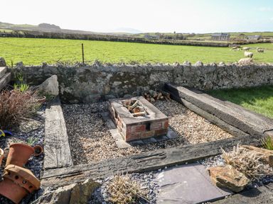 An outdoor fire pit with logs on gravel surrounded by wooden beams and a stone wall with a green field and sheep beyond at Bryn Mor in Church Bay