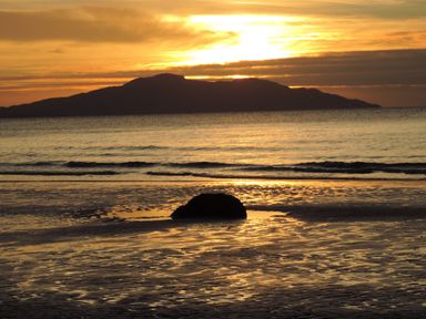 A beach at sunset with a large rock on the wet sand and an island silhouette in the background at Bryn Mor in Church Bay
